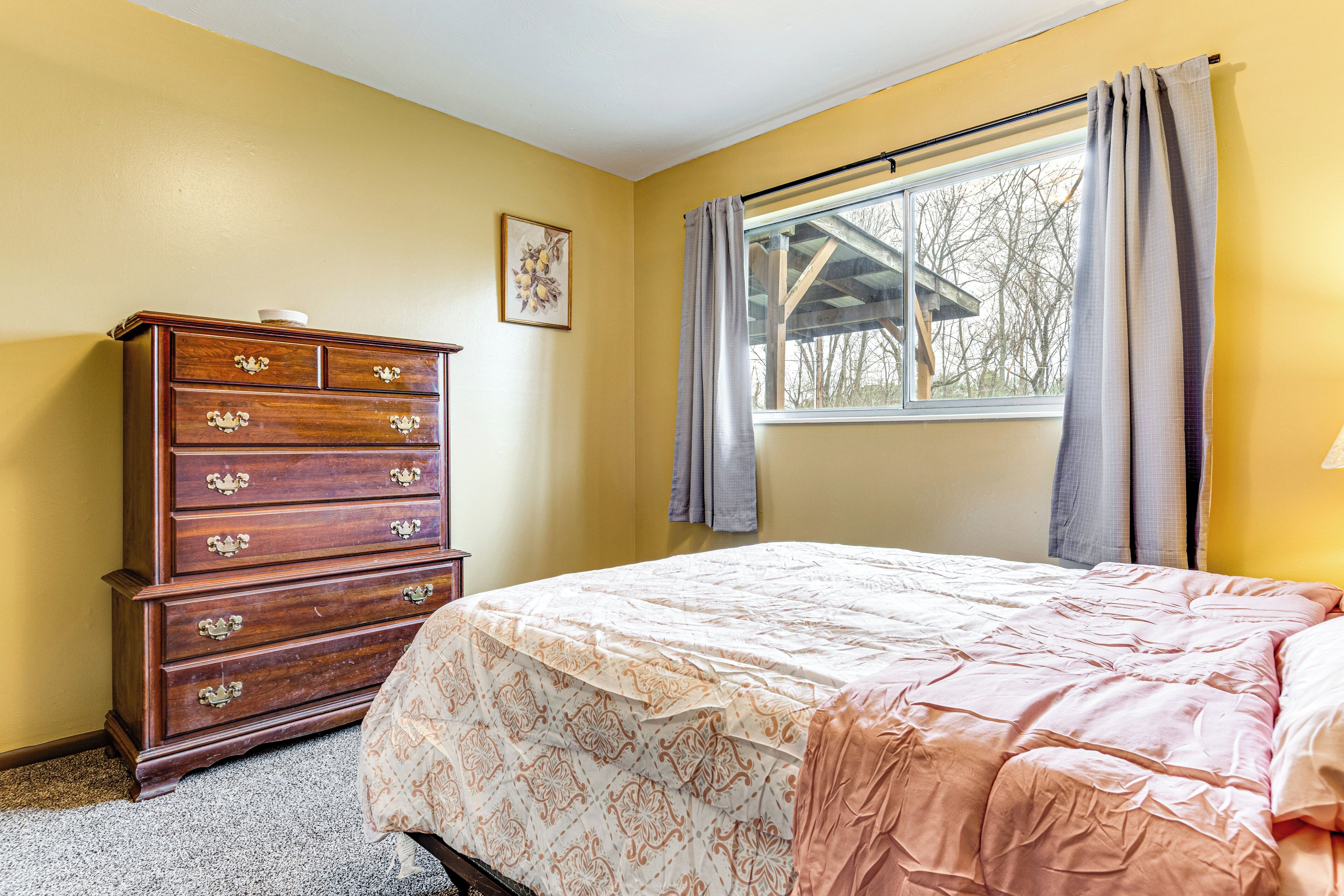 A bedroom with a wooden dresser, a bed with a patterned bedspread, a window with curtains, and a picture on the wall.