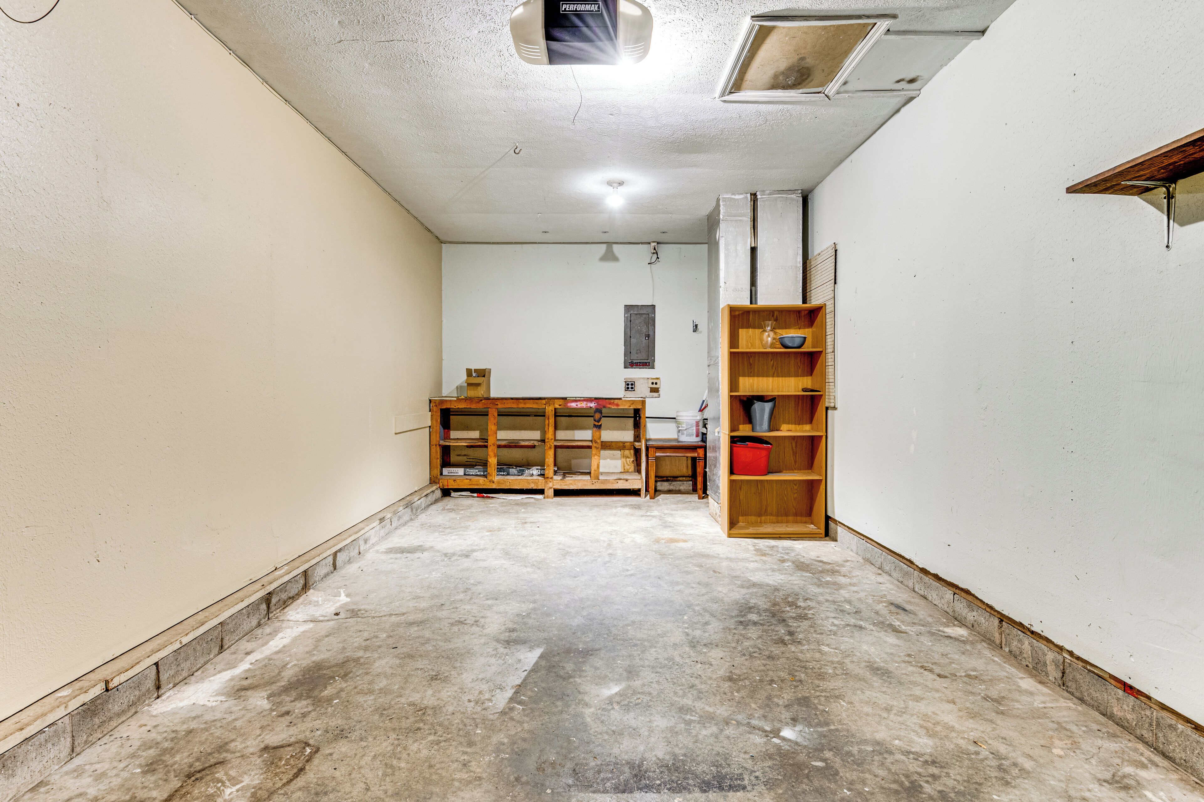 Une pièce avec un sol en béton, une étagère en bois et un mur blanc percé d’une petite fenêtre.