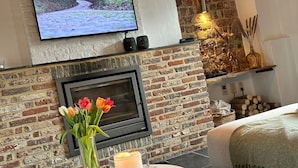 A cozy room with a fireplace, a television mounted above it, and a table with candles and flowers. - Renovated, charming heritage farmhouse in the heart of the Flemish Ardennes (Oudenaarde)
