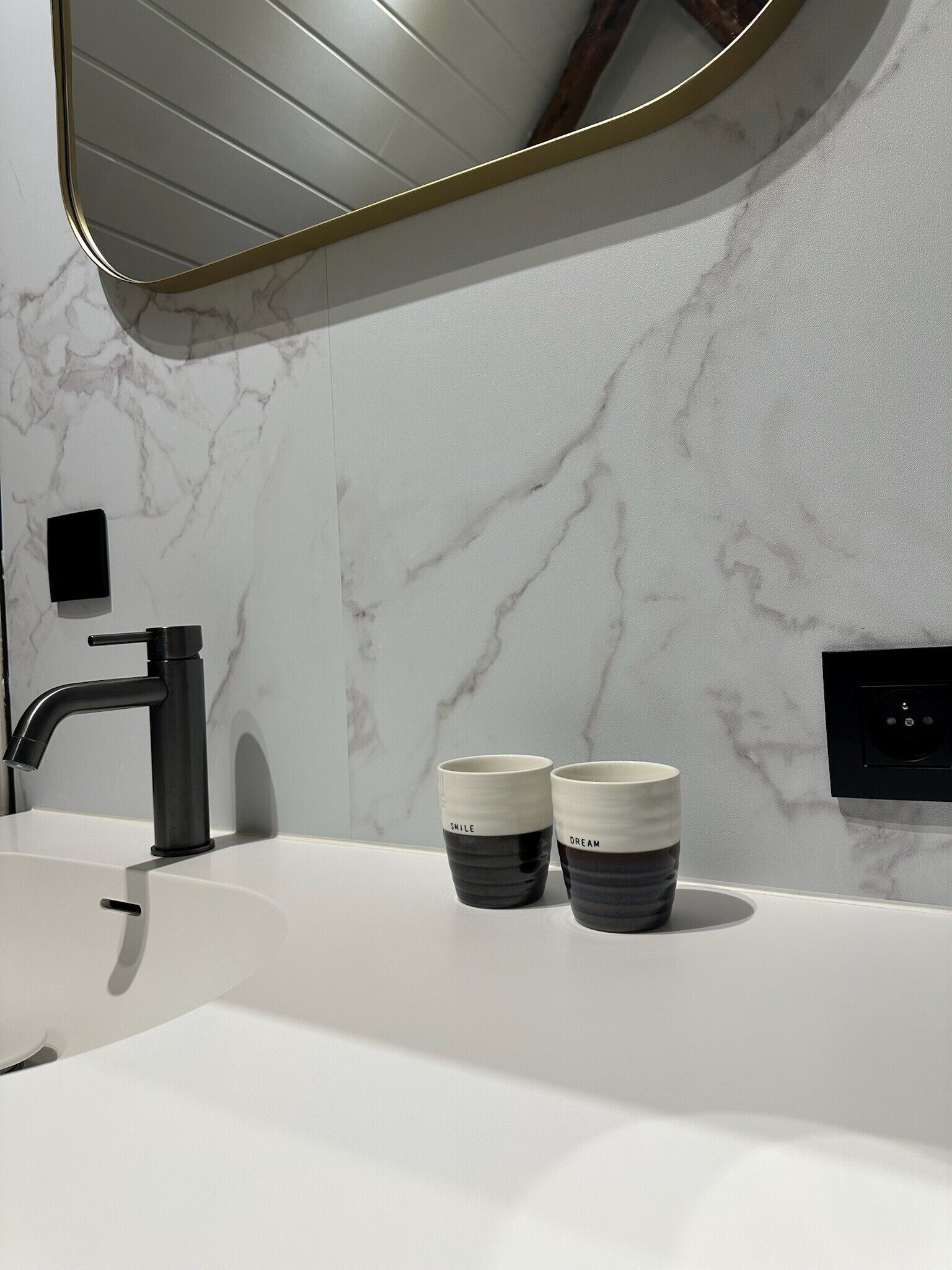 Un baño moderno con pared de mármol blanco, un grifo negro y dos tazas de cerámica con las etiquetas 'SMILE' y 'DREAM' sobre el mostrador.