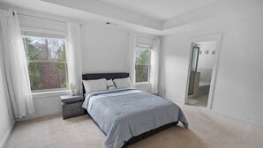 Un dormitorio con una cama, dos ventanas, una mesita de noche y una puerta que da a un baño.