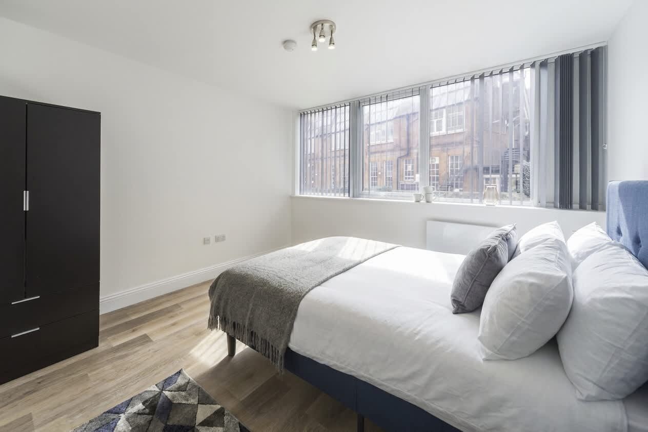 Een slaapkamer met een bed, een kledingkast, een raam met gordijnen en uitzicht op gebouwen.