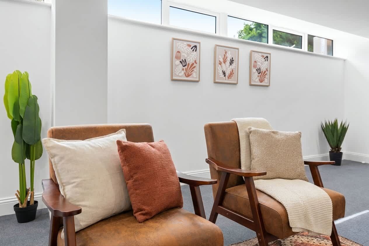 Een kamer met twee leren fauteuils, waarvan er een een beige kussen heeft en de andere een bruin kussen. Er hangen drie kunstwerken in lijsten aan de muur, en links staat een potplant.