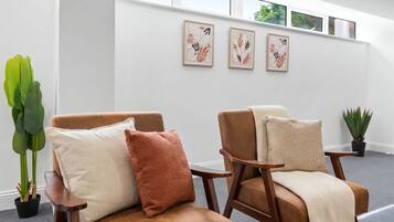 Een kamer met twee leren fauteuils, waarvan er een een beige kussen heeft en de andere een bruin kussen. Er hangen drie kunstwerken in lijsten aan de muur, en links staat een potplant.