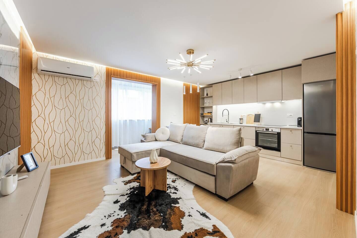 A modern living room with a sofa, a round wooden coffee table, and a kitchen area with a refrigerator, oven, and sink.