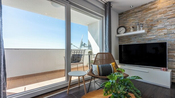 Ein modernes Wohnzimmer mit großem Fernseher, einem Couchtisch mit Pflanze, einem Korbstuhl und einem Balkon mit Aussicht.