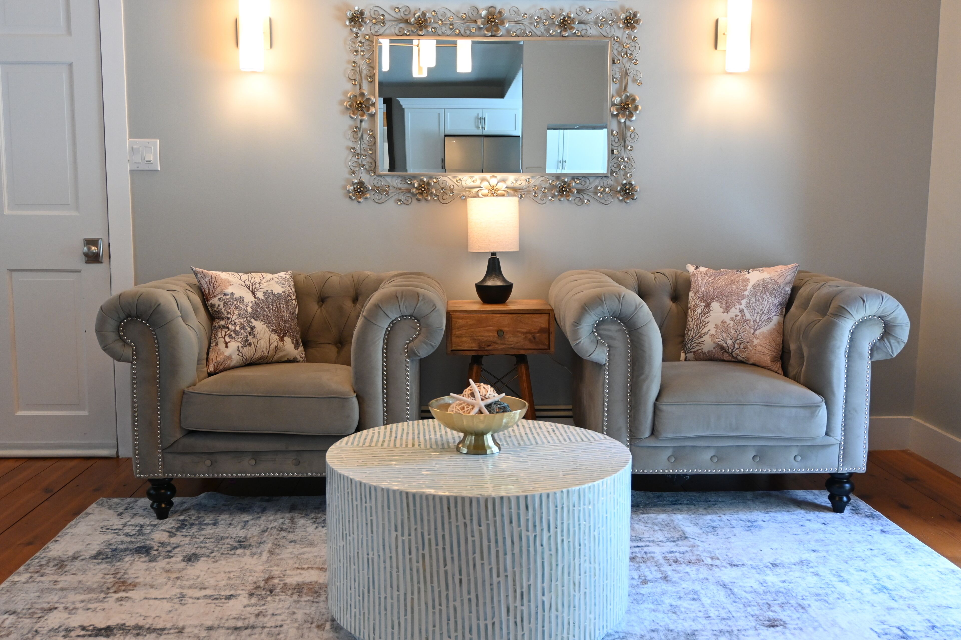 A room with two tufted sofas, a round coffee table, a mirror with a decorative frame, and a lamp.