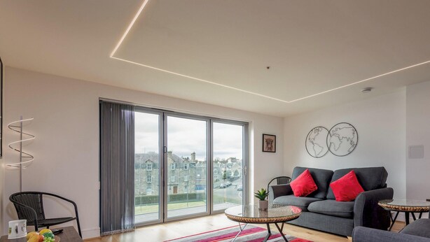 A modern living room with a sofa, a coffee table, and a large window with a view of buildings. - Square Green (No 15) | Balcony (St Andrews)