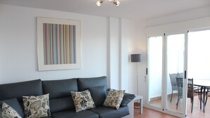 A modern living room with a grey sofa, patterned cushions, a framed artwork, a floor lamp, and a sliding glass door leading to an outdoor area with a table and chairs. - 2 Bedroom Apartment (MOJACAR)