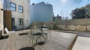 A wooden deck with outdoor furniture, a stone wall, and a modern building in the background. - FindMe inPorto Bonfim 0F (Porto)