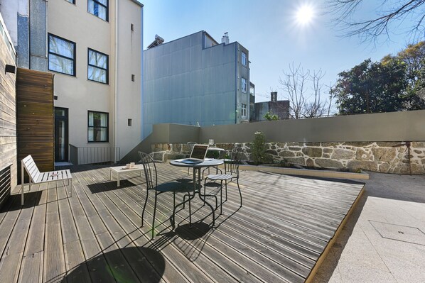 A wooden deck with outdoor furniture, a stone wall, and a modern building in the background.