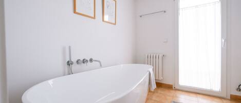 A bathroom with a freestanding bathtub, a towel rack, a radiator, and a window with blinds.