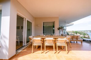 Eine sonnendurchflutete Terrasse mit einem Holztisch und Stühlen, einer Schiebeglastür und Blick auf die Küste.