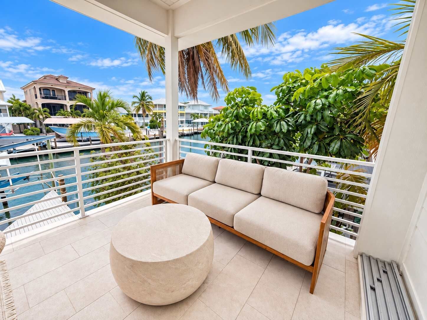 A balcony with a beige sofa and round ottoman, overlooking a marina with boats and a building in the background.