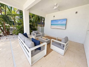 A modern outdoor living area with a sofa, chairs, a fire pit, and a ceiling fan. - Drift House · 2026 Luxury 6 King Suites Boat Dock (Fort Myers Beach)