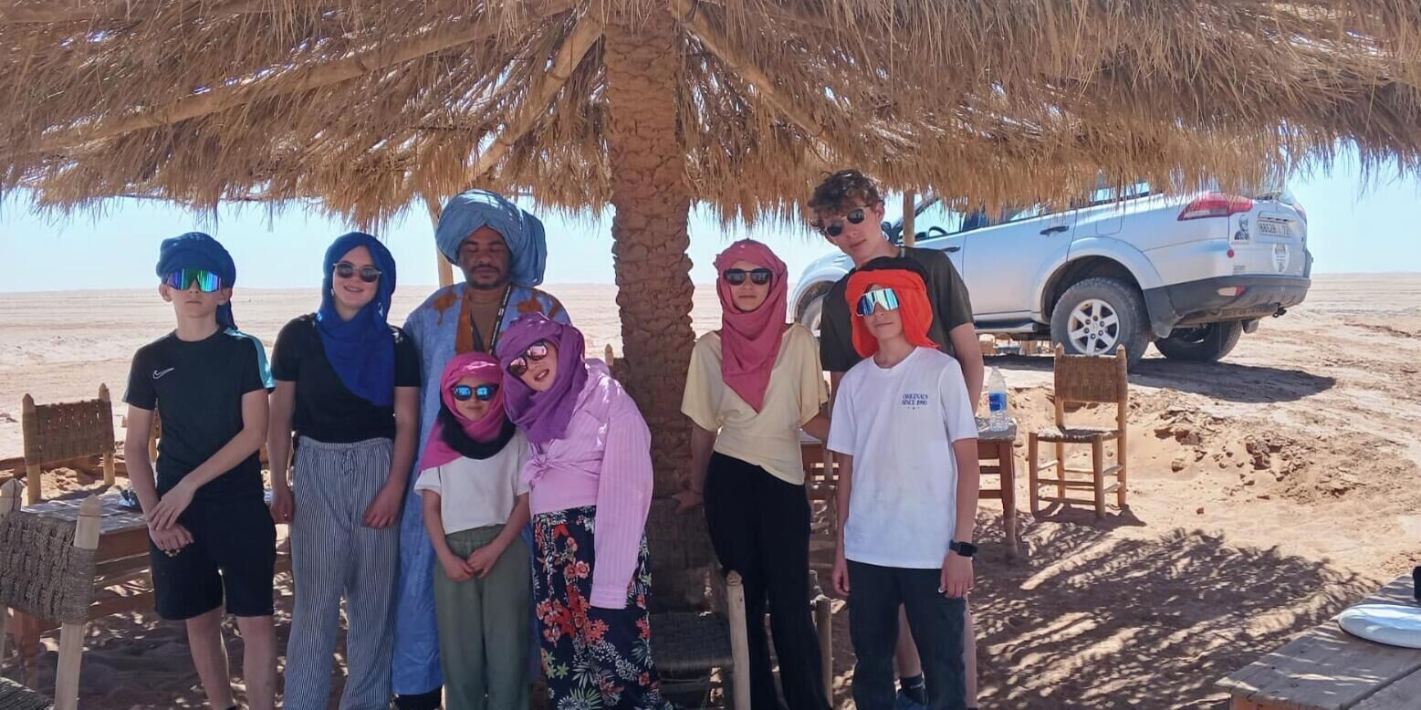 Un groupe de personnes se tient sous un palmier sur une plage de sable, avec un véhicule visible au loin.