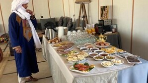 A dining area with a long table set with various dishes, a person in traditional attire, and a modern lamp. - starlitdesertcamp (Foum Zguid)