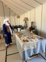 Un comedor con una mesa larga dispuesta con diversos platos, una persona vestida con traje tradicional y una lámpara moderna.
