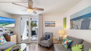 Un soggiorno con divano, poltrona e televisore. C'è una ventola a soffitto, un quadro raffigurante una spiaggia e vista sull'oceano attraverso la porta in vetro.