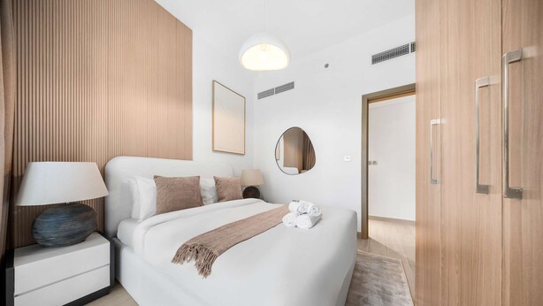 Ett modernt hotellrum med en snyggt bäddad säng, ett sängbord med en lampa, en rund spegel och en garderob.