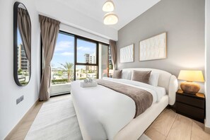 A hotel room with a large bed, a nightstand with a lamp, a window with curtains, and artwork on the wall. - Free Housekeeping Visits During Your Stay - Stayshort - Contemporary 2 Bed Retreat in Meydan for 6 (Dubai)