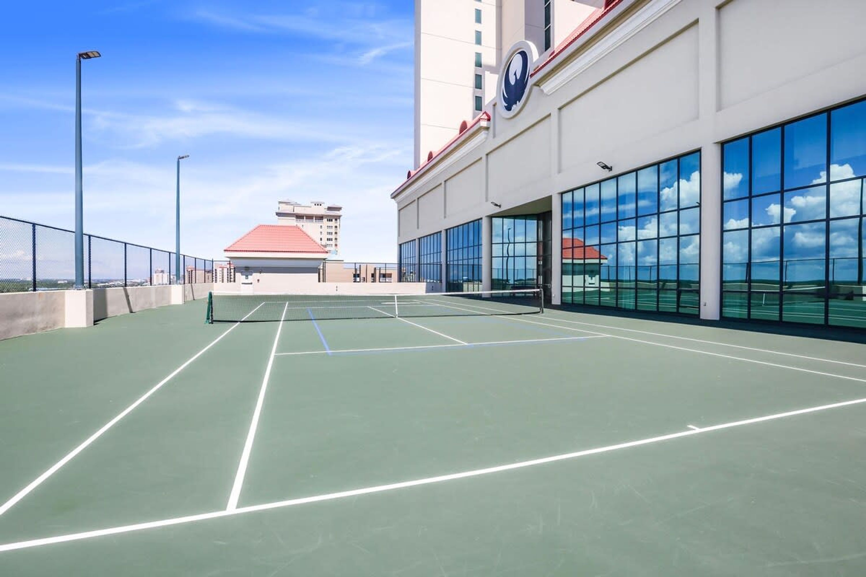 An indoor tennis court with a net, surrounded by glass walls and a building with a red roof.