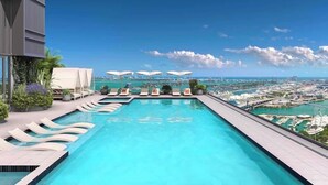 Une piscine à débordement située sur le toit, dotée de chaises longues et de parasols, offrant une vue imprenable sur le paysage urbain et l’étendue d’eau.