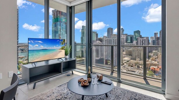 Un salon moderne avec une grande télévision, une table ronde sur laquelle sont disposées des boissons, et une vue sur la ville grâce à de grandes fenêtres.