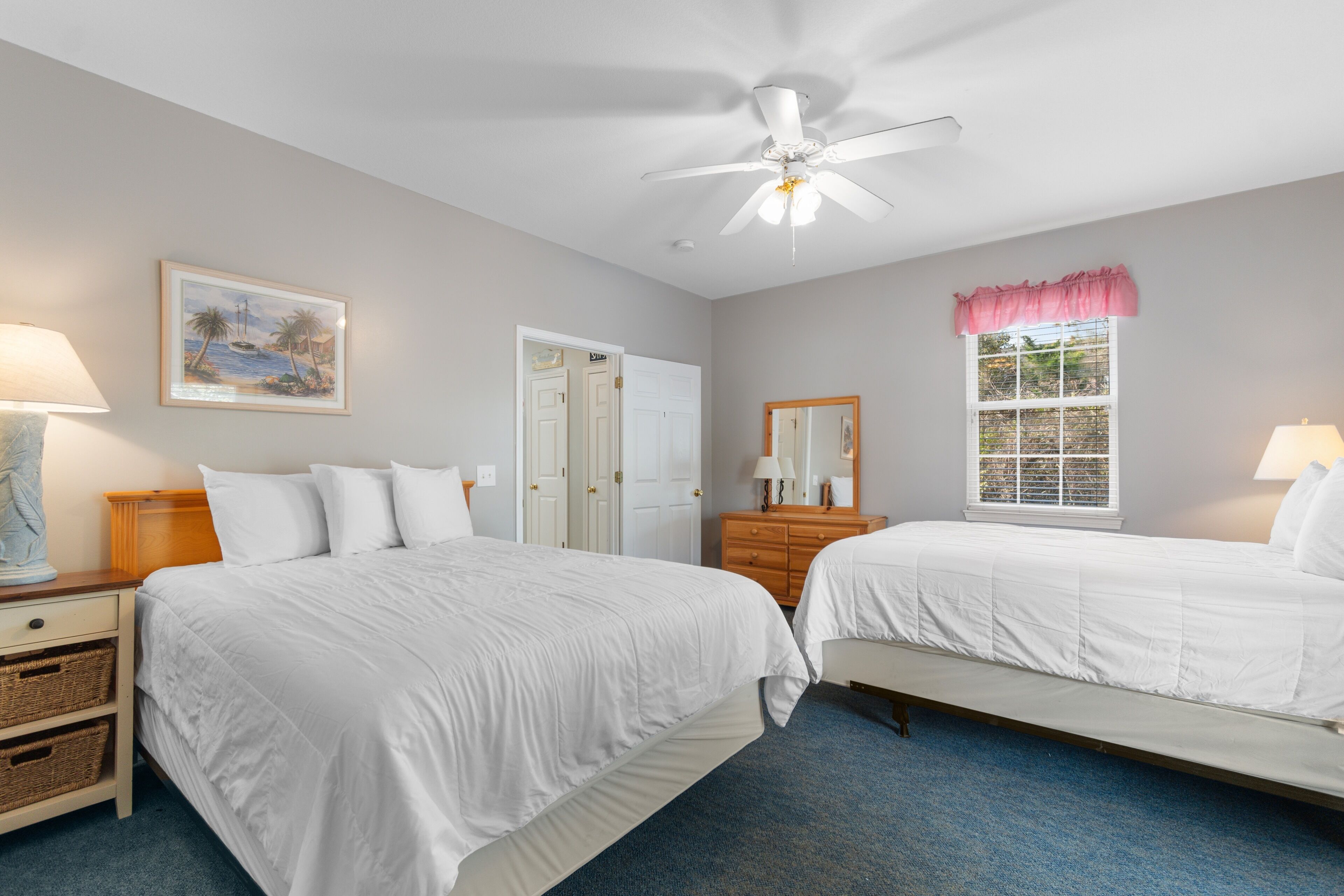 A hotel room with two beds, a ceiling fan, a painting, a window with a pink valance, and a wooden dresser.