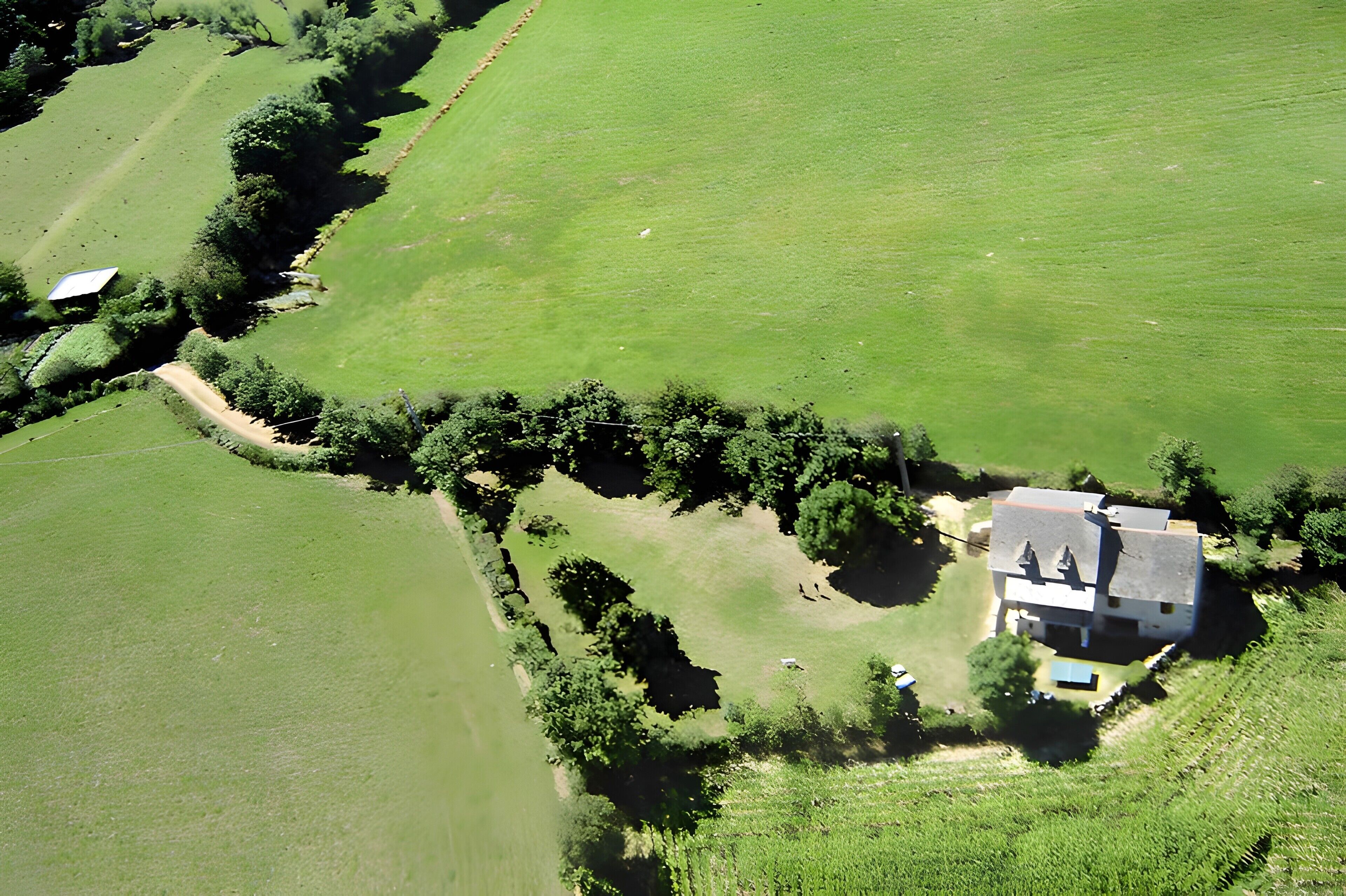 Vue aérienne d’une propriété rurale comprenant une maison, un espace clôturé et des champs environnants.