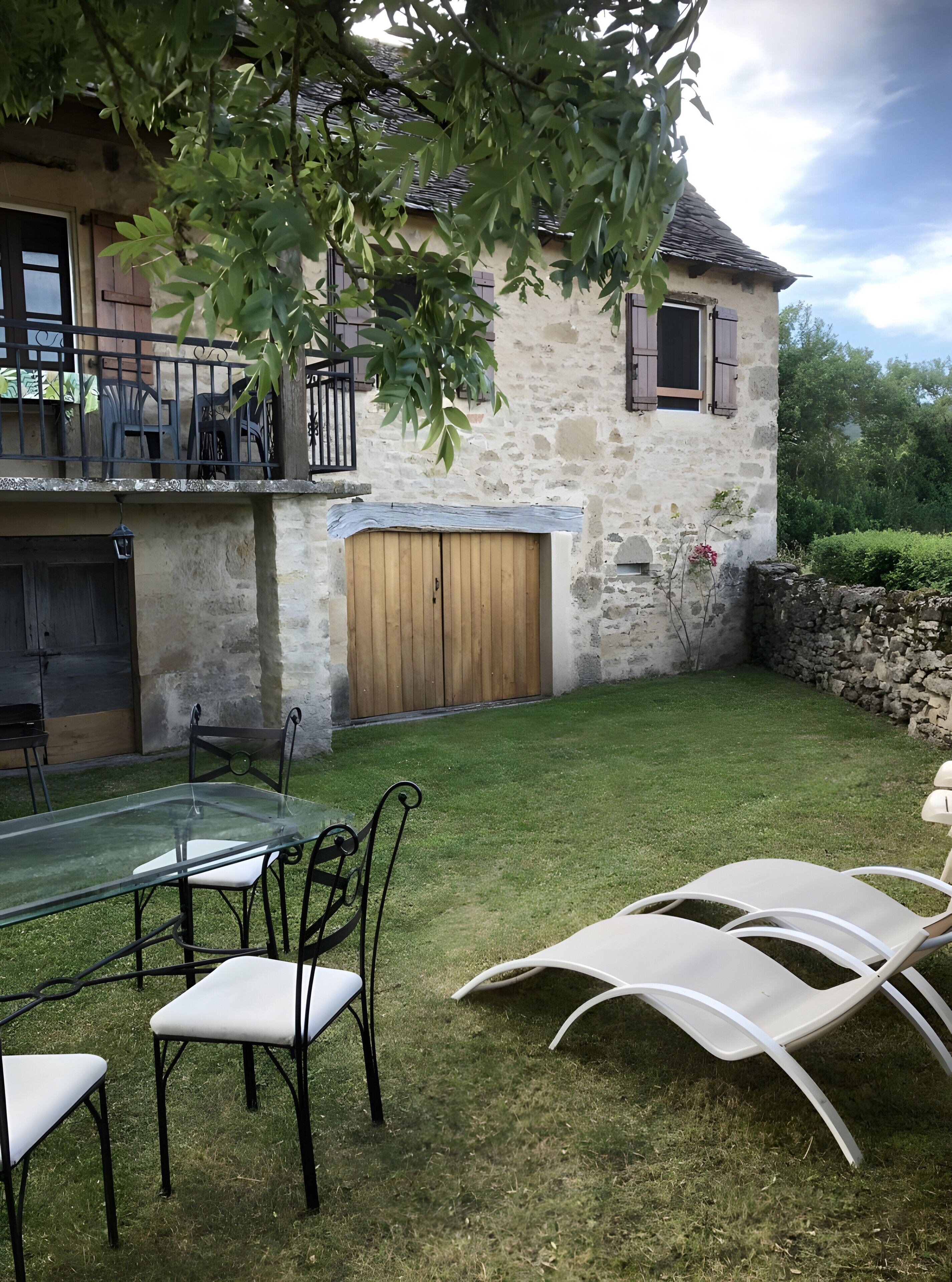 A stone house with a wooden door, a balcony with chairs, and a grassy yard with outdoor furniture.