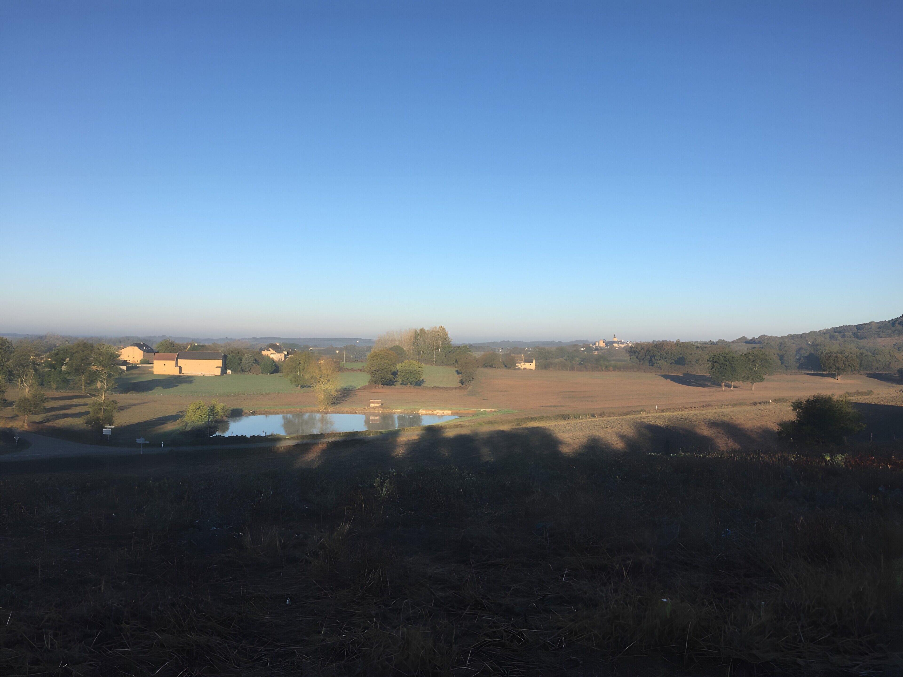 Un paysage rural avec des champs, un étang et des bâtiments situés au loin.