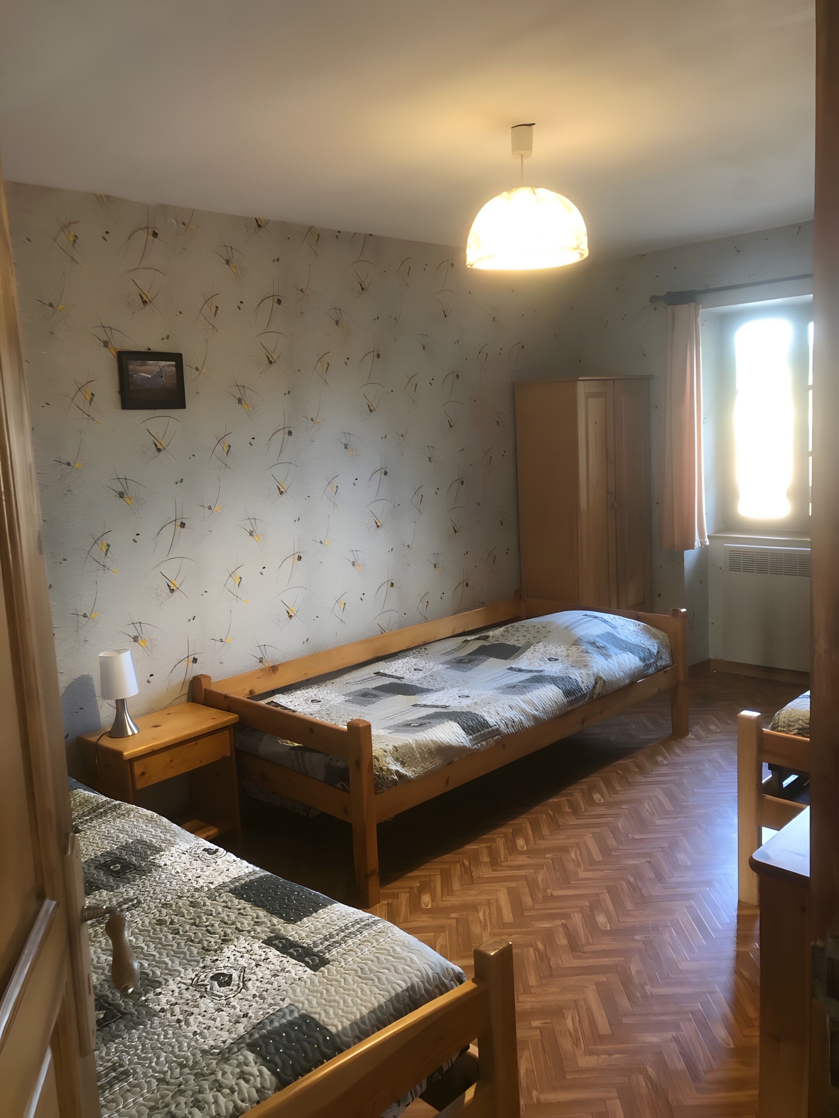 A small, cozy room with a bunk bed, a wooden wardrobe, a bedside table with a lamp, and a patterned rug.