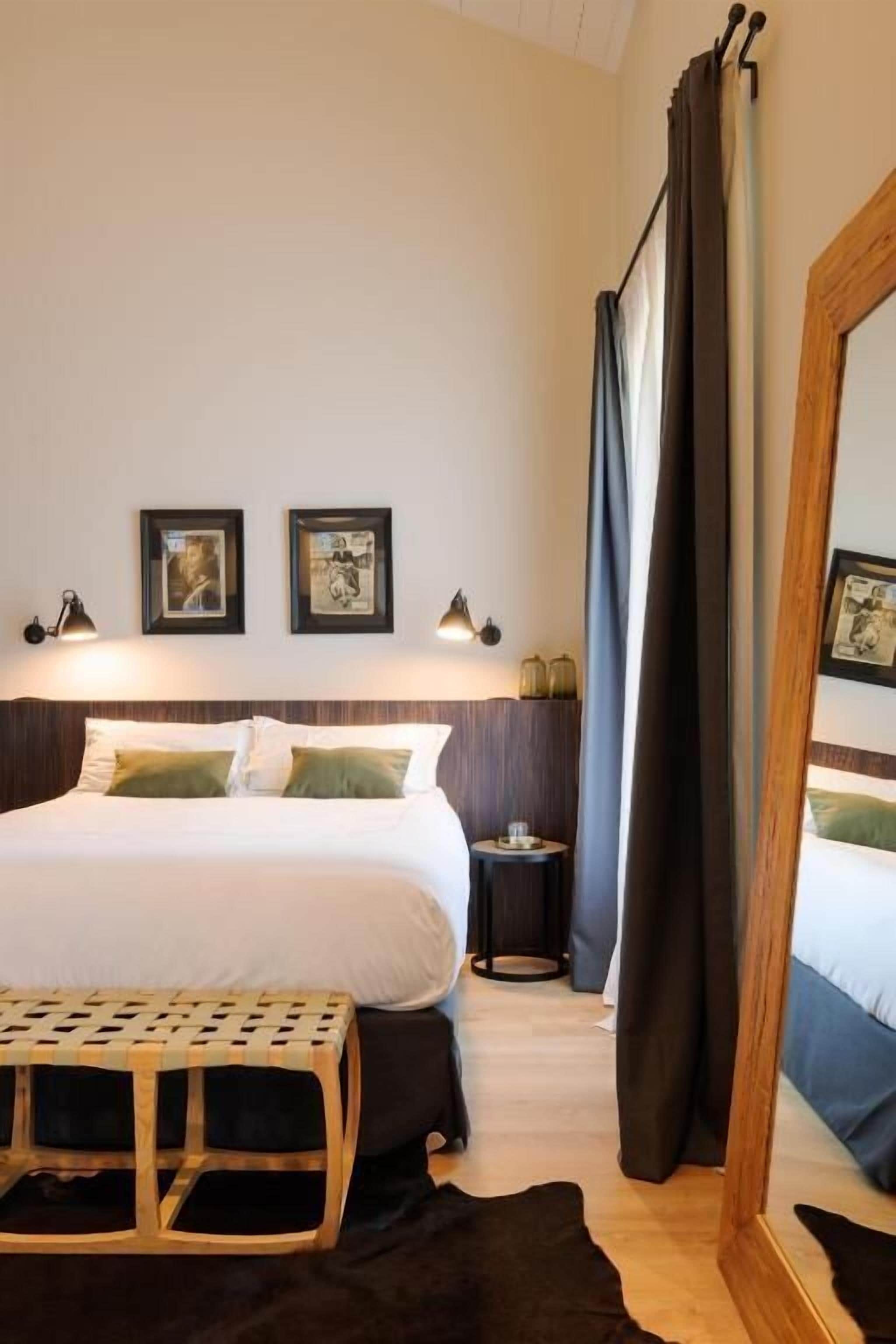 A hotel room with a bed, a small table, a mirror, and framed pictures on the wall.