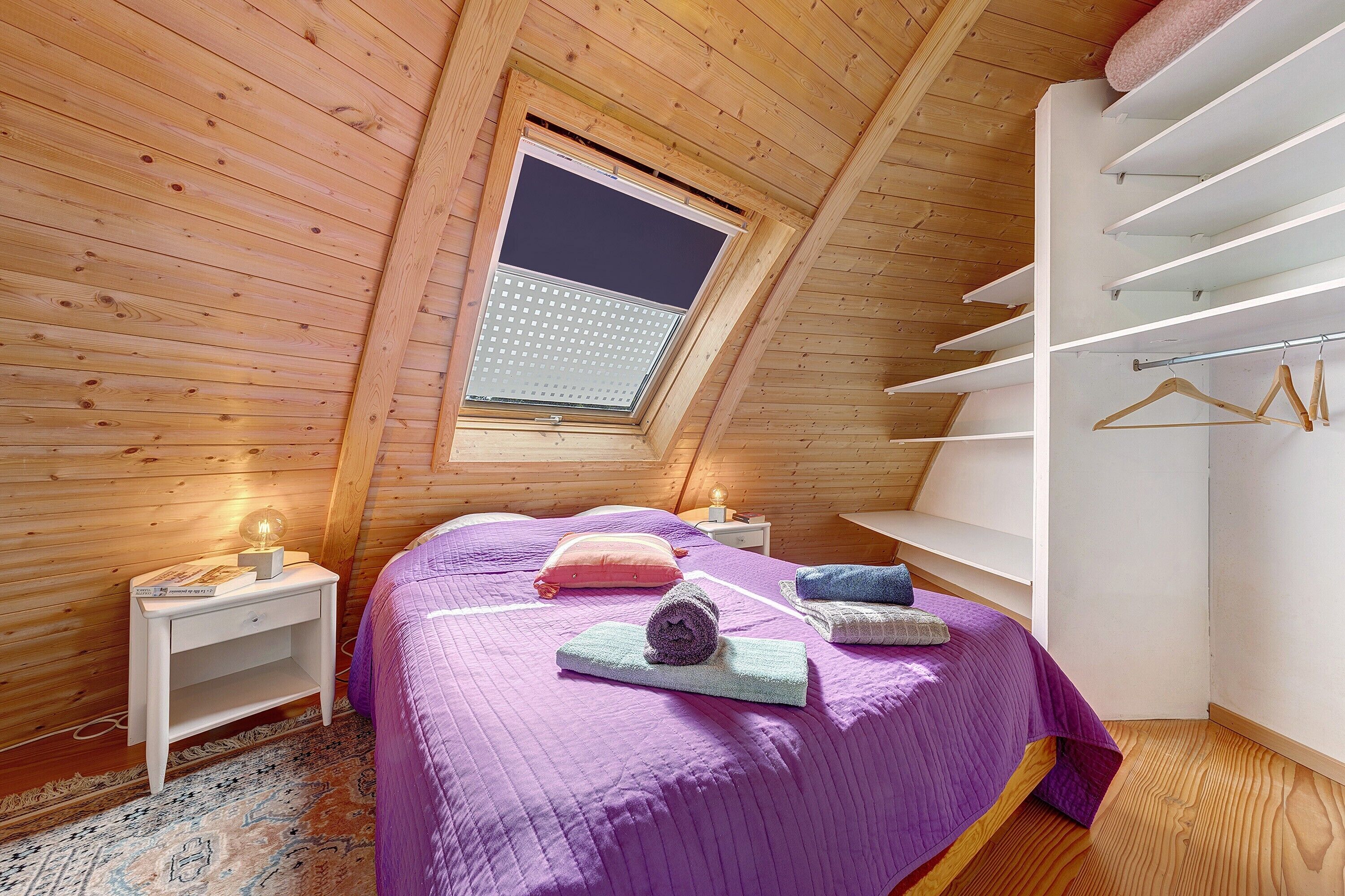 Une chambre avec un plafond en pente, un puits de lumière, un lit avec une literie violette, une table de chevet avec une lampe et une armoire intégrée avec un espace pour suspendre les vêtements.