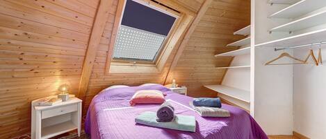 Une chambre avec un plafond en pente, un puits de lumière, un lit avec une literie violette, une table de chevet avec une lampe et une armoire intégrée avec un espace pour suspendre les vêtements.