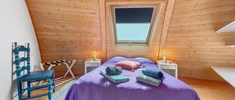 A bedroom with a sloped ceiling, wooden walls, a window, a bed with a purple bedspread, two bedside tables with lamps, and a chair.
