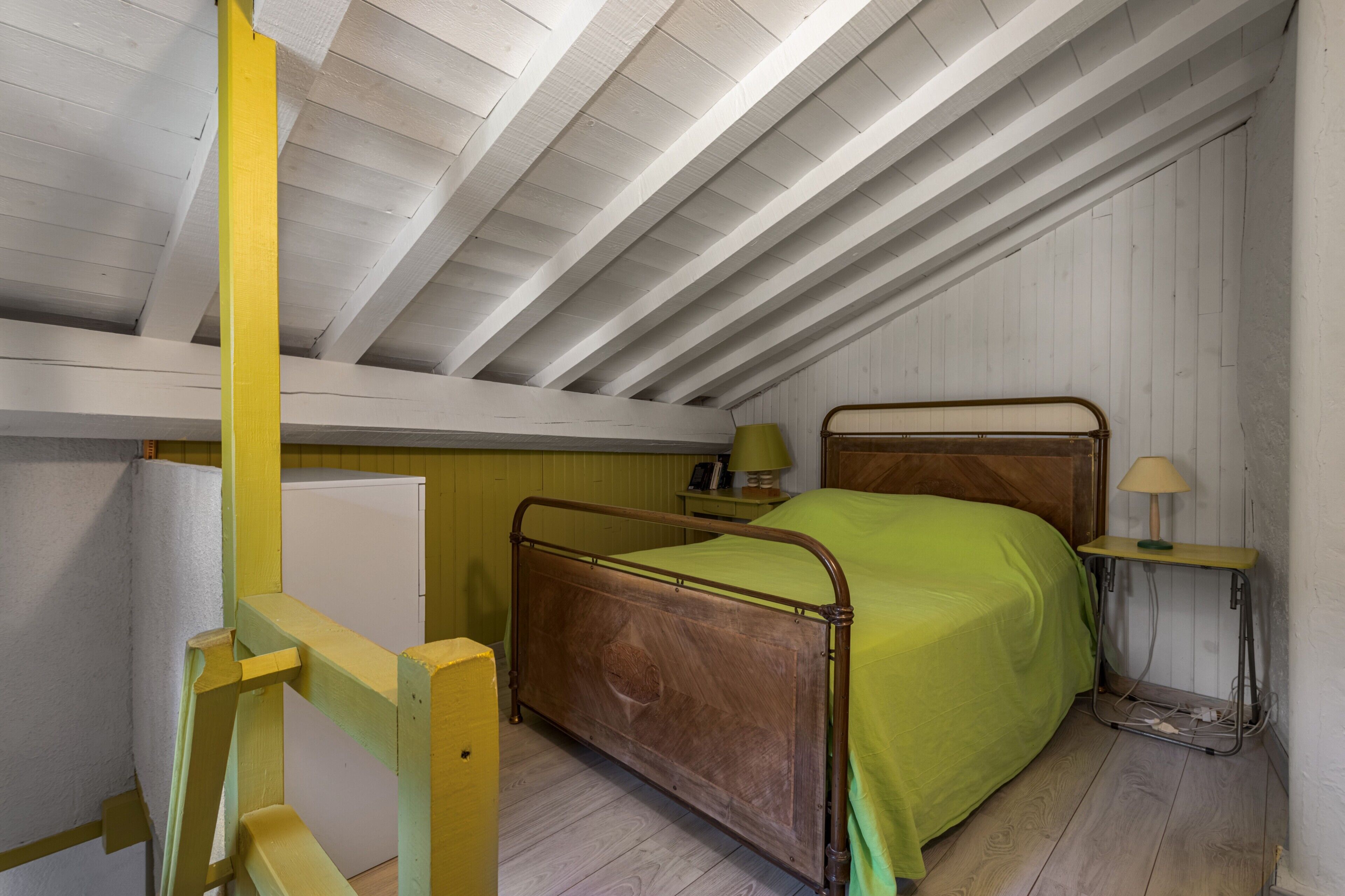 Un lit simple avec un cadre en bois et une couvre-lit verte, dans une pièce aux murs en bois peints en blanc et au plafond en pente.
