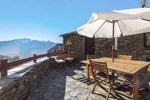 Un bâtiment en pierre avec une table et des chaises en bois, un parasol blanc et une vue sur les montagnes.