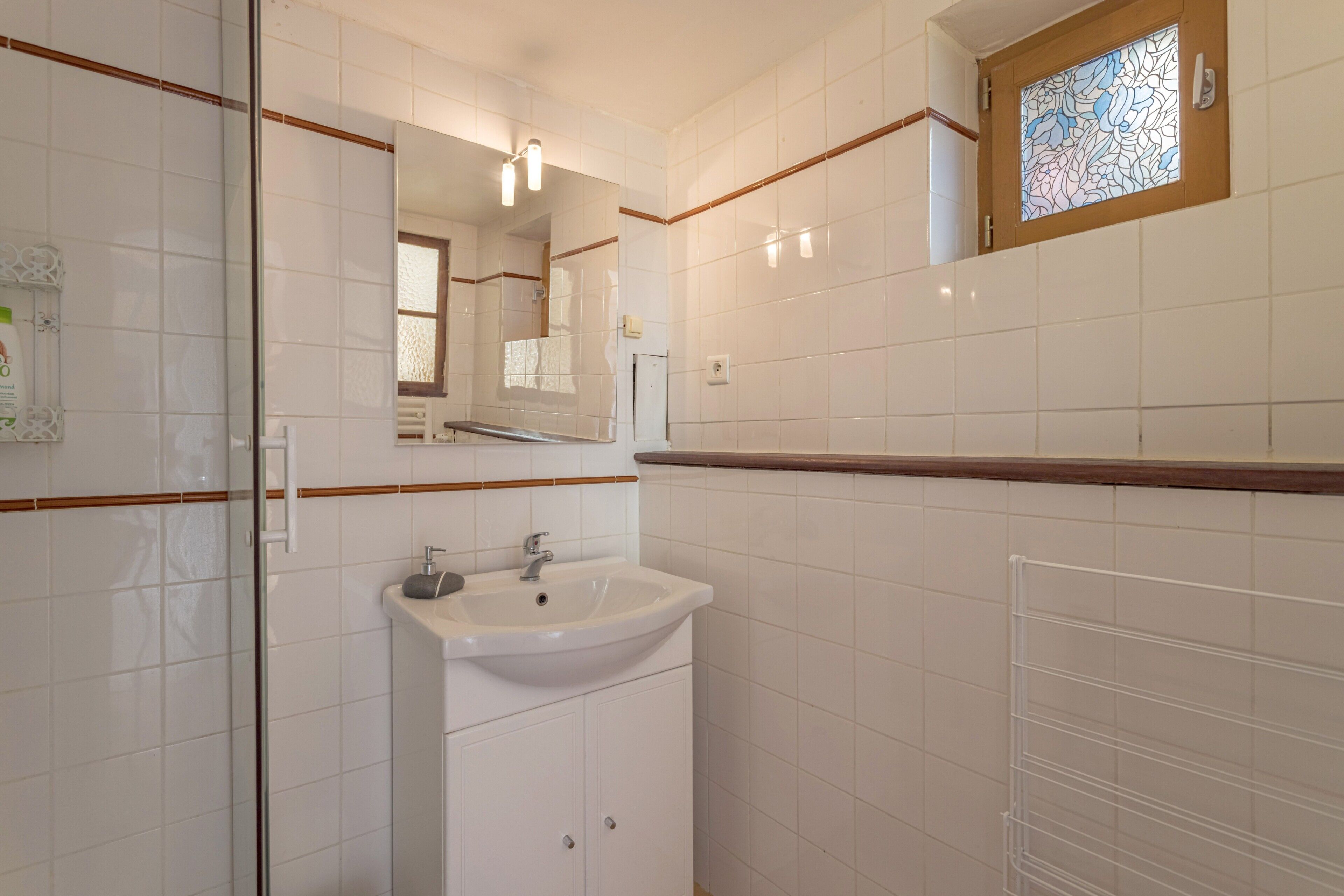 A bathroom with white tiles, a sink, a mirror, and a window with stained glass.