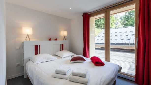 A hotel room with a bed, two bedside lamps, a window with red curtains, and a view of a building outside. - Chalet in Paradiski With Spa & Stunning Views (Peisey-Nancroix)