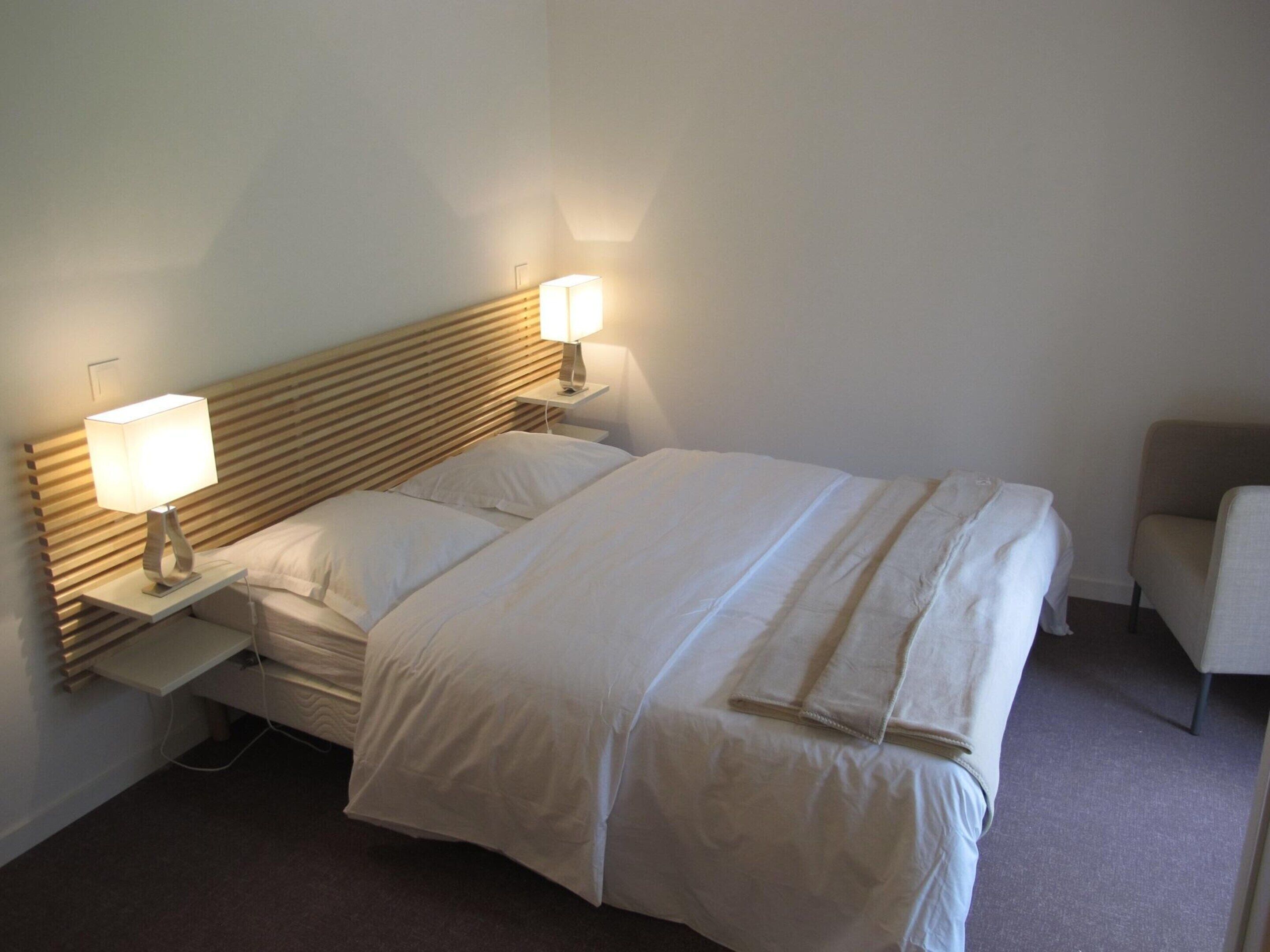 A neatly made bed with white linens, two bedside lamps, a wooden headboard, and a small chair in a simple room.