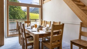 A dining area with a wooden table and chairs, a view of a mountain landscape through large windows, and a wooden staircase. - Chalet in Paradiski With Spa & Stunning Views (Peisey-Nancroix)
