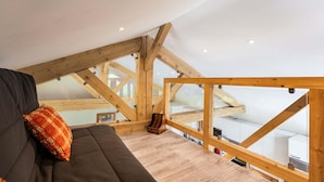 A loft bedroom with a sloped ceiling, wooden beams, a dark sofa, and a patterned pillow. - Chalet in Paradiski With Spa & Stunning Views (Peisey-Nancroix)