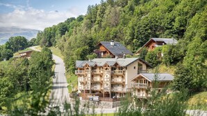 A multi-story wooden lodge with balconies, surrounded by lush greenery and a winding road. - Chalet in Paradiski With Spa & Stunning Views (Peisey-Nancroix)
