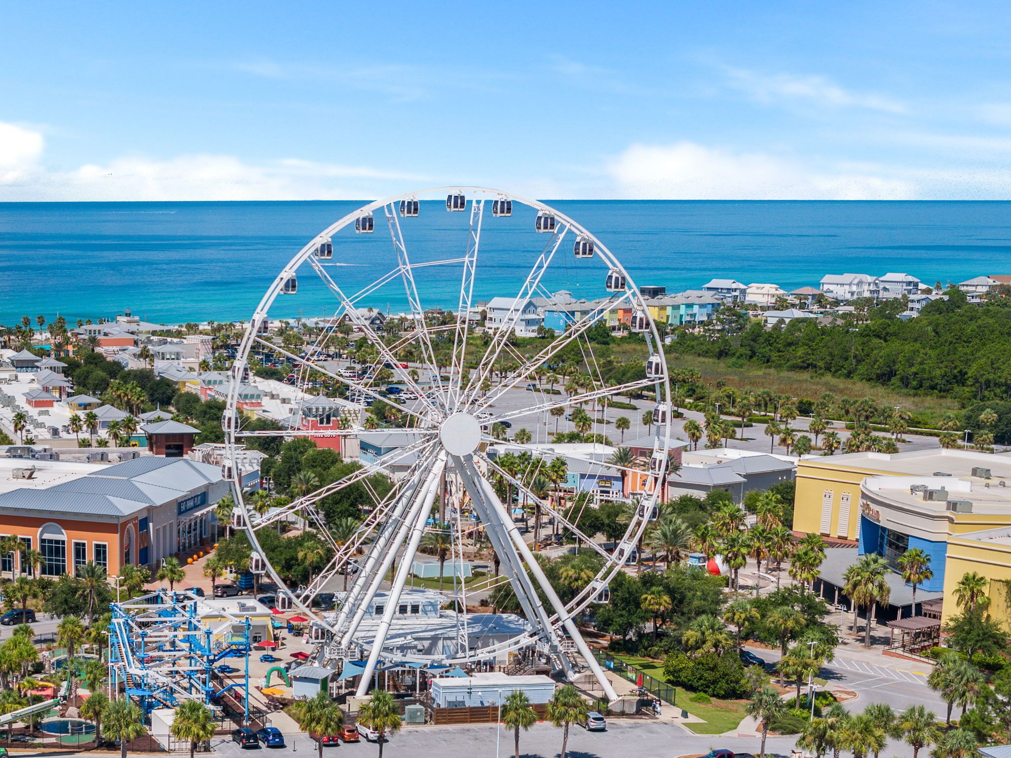 Une grande roue avec des cabines, un parking et une vue sur l’océan et les bâtiments.