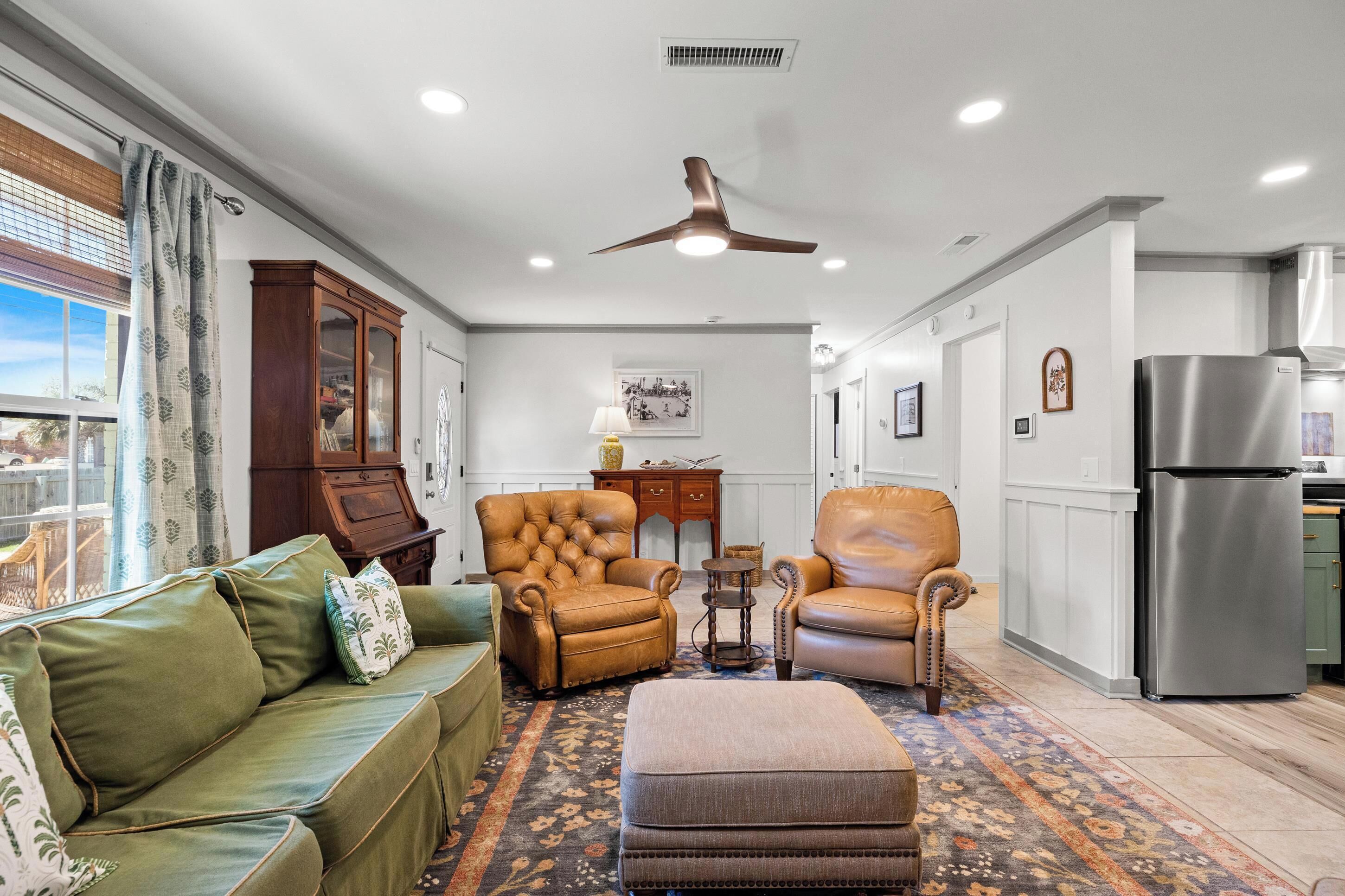 A living room with a green sofa, two leather armchairs, a patterned rug, a ceiling fan, and a kitchen with a stainless steel refrigerator.