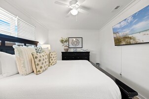 A bedroom with a bed, a ceiling fan, a dresser, a framed picture, and a TV on the wall.