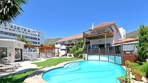 Ein Schwimmbad mit einer Terrassenfläche, einem überdachten Sitzbereich und einem Gebäude mit Balkon.