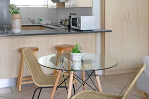 A modern kitchen with a glass-top table, wicker chairs, a microwave, and a potted plant. - Sea Point Studio Pool Gym BBQ (Cape Town)